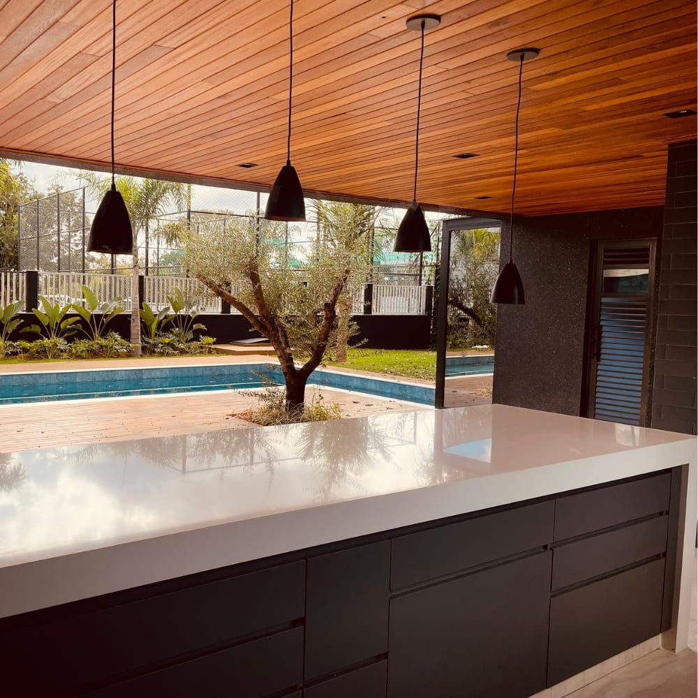 Ambiente moderno com forro de madeira Angelim Extra instalado no teto, integrando área gourmet com bancada de pedra branca, pendentes pretos e vista para piscina com deck. O Forro Angelim Extra destaca-se pela tonalidade quente e acabamento sofisticado.