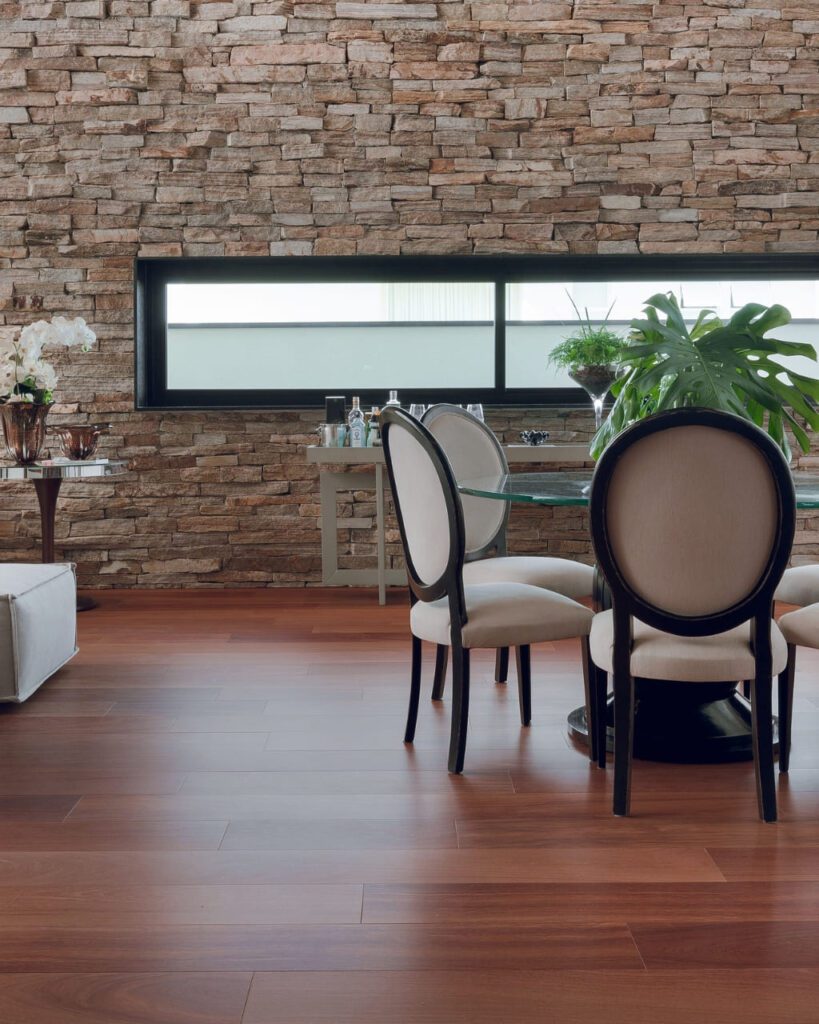 Ambiente sofisticado com piso de madeira nobre em tom avermelhado, parede de pedras e mesa de jantar elegante com cadeiras estofadas. O piso destaca o brilho e a textura natural da melhor madeira, trazendo aconchego e elegância ao espaço.